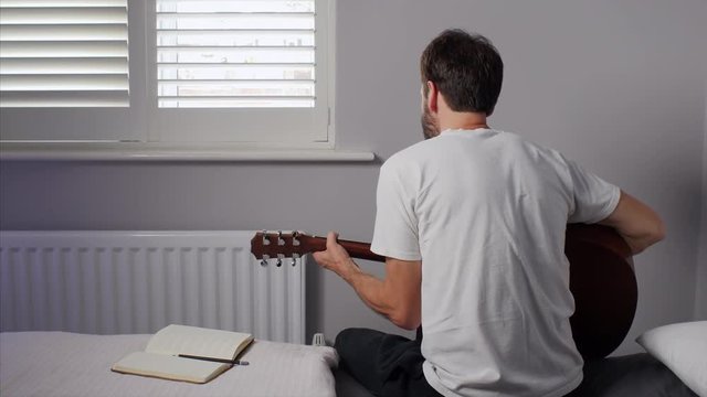 A STATIC SHOT of an average man playing his acoustic guitar and singing with passion, facing away from the camera, in a minimalistic bedroom.