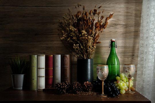 still life of wine glass and wine bottle on wooden table with grapes and a vase of dry flowers and books