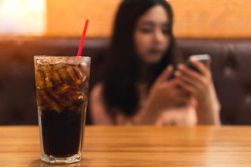 Close up glass of soft drink cola with ice on wooden table with blurred of young asian woman using smart phone.