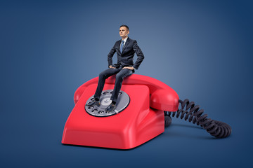 A tiny serious businessman sits patiently on a giant red retro phone with a dial.