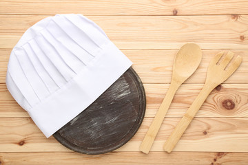 Chef hat with cooking cutlery and cutting board on brown wooden table