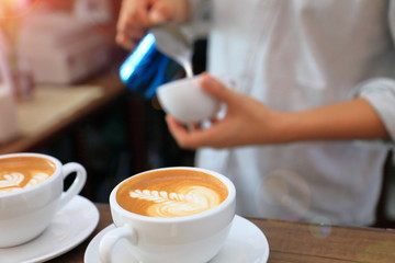 Barista using pitcher for pouring milk to cup of coffee latte with finished cup on the foreground in the cafe