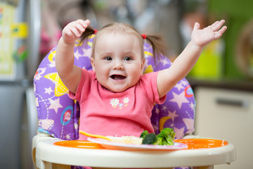 Fototapeta premium Joyful baby eats pasta and broccoli sitting in highchair in kitchen