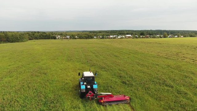 Sky view of slowly moving big blue tractor with attached rectangular red mower deck and mowing large yellow and green surface of field surrounded by trees.
