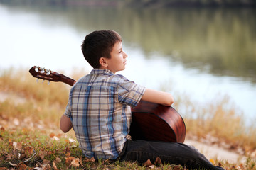 the boy plays an acoustic guitar, sits on the Bank of the river, autumn forest at sunset, beautiful nature and the reflection of trees in the water