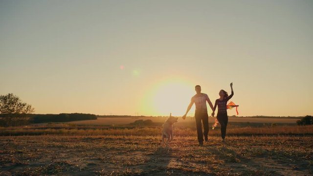 Beauty young couple with dog running with kite on the field. Beautiful family with flying colorful kite over sunset sky. Free, freedom concept. Emotions, healthy lifestyle.