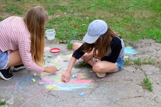 Two teen girls  with long hair wearing shorts drawing with colorful chalk crayons on old grunge cracked concrete  outdoors in yard on green grass background in summer day. Children creative outdoors a