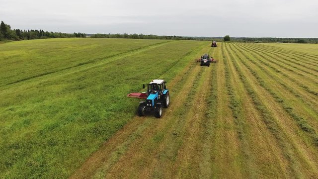 Camera shows three big equipped tractors with hay mower, hay baler and raker moving one by one on large yellow and green farmland field surrounded by trees.