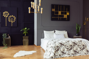 Gold chandelier above bed in grey bedroom interior with black posters and plants. Real photo