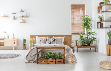 Bed with wooden headboard in white spacious bedroom interior with cupboard and plants. Real photo