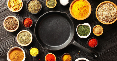 From above view of black pan placed in middle with many bowls of spices around on wooden background 