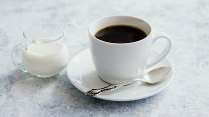 Fototapeta na wymiar Arrangement of small glass pitcher with fresh milk on marble table with white cup of coffee with silver spoon on saucer
