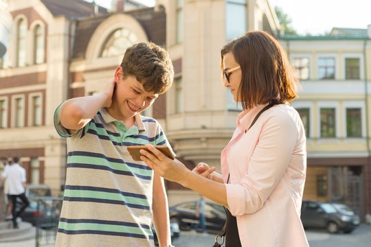 Parent and teenager, relationship. Mother shows her son something in the mobile phone, boy is embarrassed, smiling, holding his hands on his head, city street background