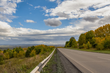 The highway along the beautiful autumn trees leaves into the distance, in the blue sky large clouds.