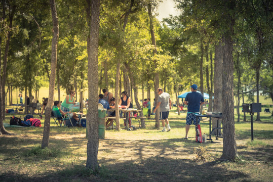 Blurred Large Group Of Friend, Family Members Enjoy Barbecue On Lakeside Area. Outdoor Camping At Natural Park With Picnic Table In Grand Prairie, Texas, USA. Outside Party And BBQ Concept