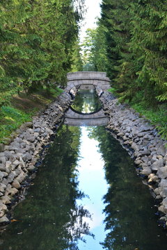 Old round stone bridge in the coniferous forest over a long narrow ditch with blue water between the stone walls in a beautiful city garden