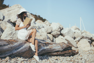 Young beautiful dreaming woman sitting alone on the trunk of a fallen tree on the beach and reading book, wearing white dress, hat and shoes - relaxing and traveling concept - romantic italian seaside