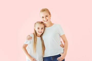 smiling mother and daughter in white t-shirts embracing and looking at camera isolated on pink