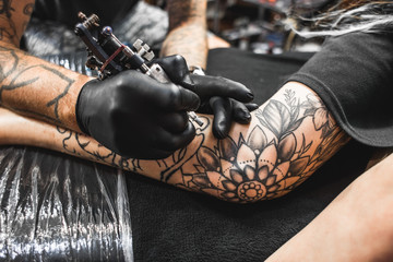 girl with dreadlocks in a tattoo parlor. The master creates a picture on the body of a young beautiful girl. Close-up of hands and tattoo machine - obrazy, fototapety, plakaty