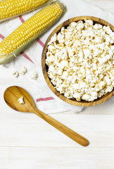 Brown wooden bowl with delicious traditional popcorn, fresh corn and a spoon on a light wooden background. Top view of a light meal background.