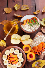 Oatmeal with different fruits and honey. Oat flakes with milk and dried apricots on wooden background. Healthy food for vegan. Dry oats with berries of mountain ash and green mint. Autumn pattern