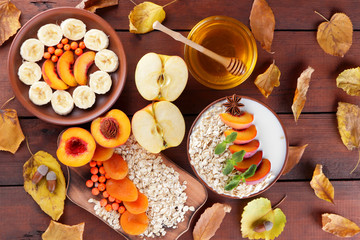 Oatmeal with different fruits and honey. Oat flakes with milk and dried apricots on wooden background. Healthy food for vegan. Dry oats with berries of mountain ash and green mint. Autumn pattern