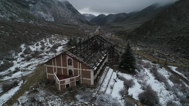 Aerial drone scene of destroyed building, old burned hotel in snowy grey day in Tunuyan river Valley. Camera moves along constructions roof metal structure. Snowy mountains and vegetation