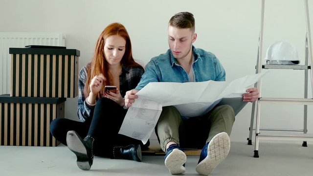 Young couple counting bills with smartphone and blueprints sitting on floor at their new home
