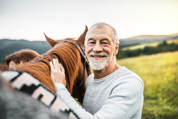 A senior man standing close to a horse outdoors in nature, holding it.