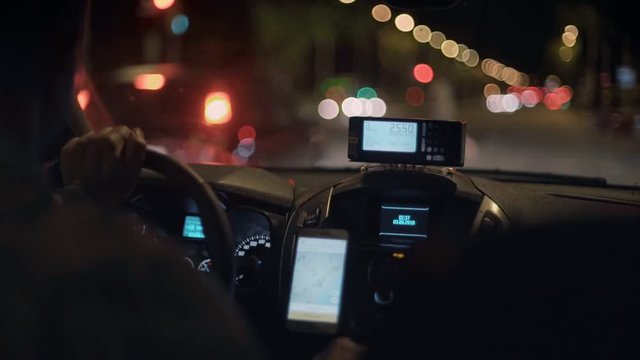Taxi driver drives cab through night city streets or highway road, uses smartphone for navigation or uses shared economy phone app to find passengers or clients, taximeter counts fare