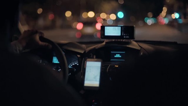 Taxi driver drives cab through night city streets or highway road, uses smartphone for navigation or uses shared economy phone app to find passengers or clients, taximeter counts fare