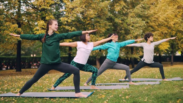 Young ladies yoga students are standing in warrior pose then moving into triangle position during outdoor class in park in autumn. Youth and health concept.
