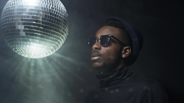 Low-key portrait shot of cool black man in hat and sunglasses rapping and spinning disco ball while partying isolated on dark background in foggy studio