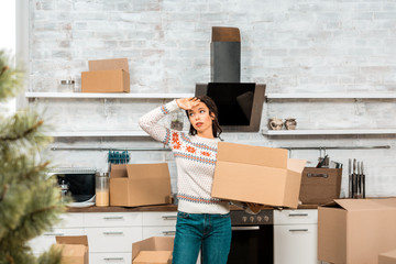 tired pretty woman holding cardboard box and wiping forehead in kitchen at new home