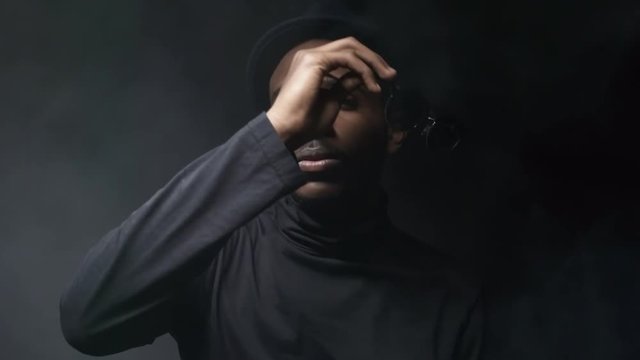Low-key portrait shot of confident black man in hat taking off sunglasses and posing isolated on dark background in foggy studio
