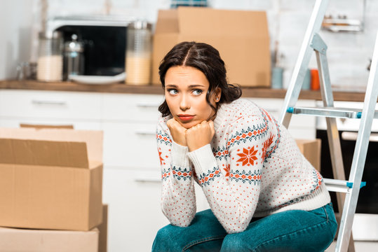 sad beautiful woman sitting on laddder in kitchen with cardboard boxes during relocation at new home