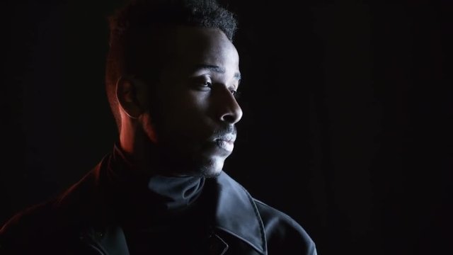 PAN low-key portrait with red lighting: serious black man in leather jacket standing isolated on dark background and looking away, then turning his head and looking at camera with calm expression