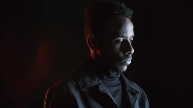Low-key portrait with red lighting: confident black man in leather jacket standing isolated on dark background and moving his head while looking away from camera