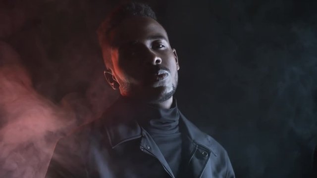Low-key portrait shot with red lighting: confident black man in jacket looking at camera and gesturing while posing in dark studio filled with smoke