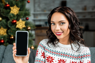 beautiful woman showing smartphone with blank screen on sofa near christmas tree at home