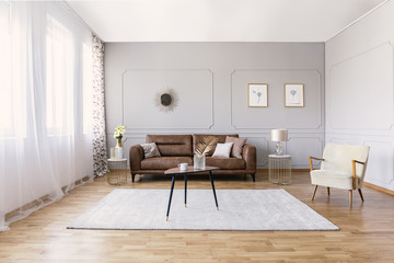 Brown leather sofa in elegant living room interior with stylish armchair, coffee table and drawings on the grey wall