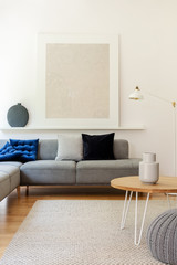 Grey poster above settee with pillows in white living room interior with wooden table. Real photo
