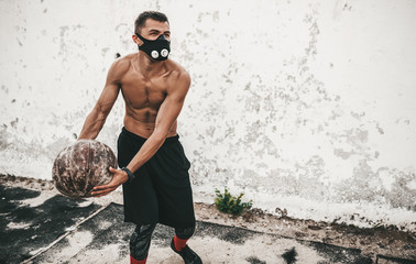Young muscular athlete male doing exercicses with medicine ball in training mask on concrete background outside with copy space for your advertising. Shirtless sportsman doing workout crossfit.