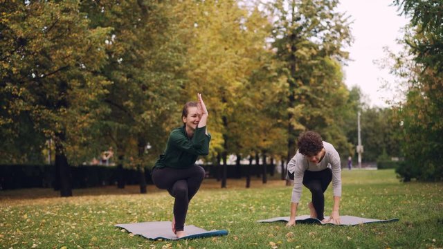 Female yoga instructor is teaching starting student Eagle pose in park, cheerful girl is making mistakes and laughing, women are enjoying nature and exercise.
