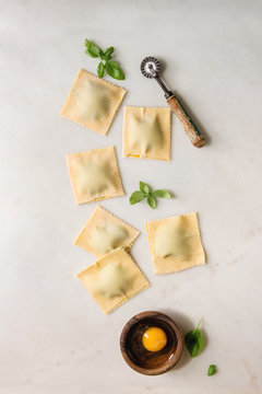 Homemade raw uncooked italian pasta ravioli staffed by spinach ricotta, egg yolk, basil, pasta cutter. White marble background. Flat lay, space