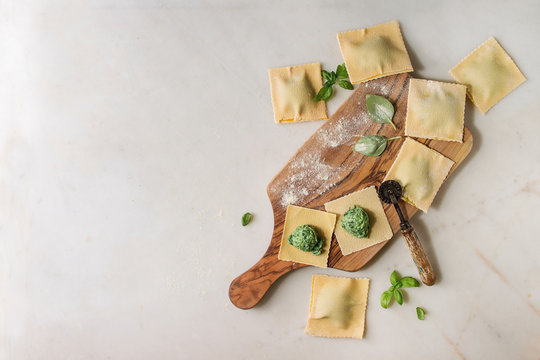 Homemade raw uncooked italian pasta ravioli staffed by spinach ricotta, egg yolk, basil on olive wood cutting board. White marble background. Flat lay, space