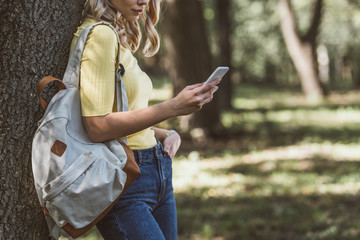 partial view of woman with backpack on shoulder using smartphone in forest