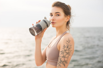 Young thoughtful woman in sporty top holding sport bottle in hand while dreamily looking in camera with beautiful sea on background