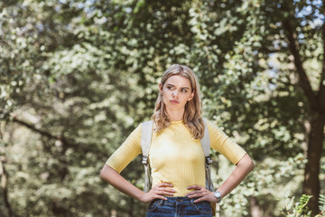 portrait of thoughtful woman with backpack standing akimbo in park