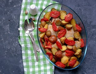 Baked chicken with potatoes and tomatoes in a glass form for baking on a dark gray background. Top view.